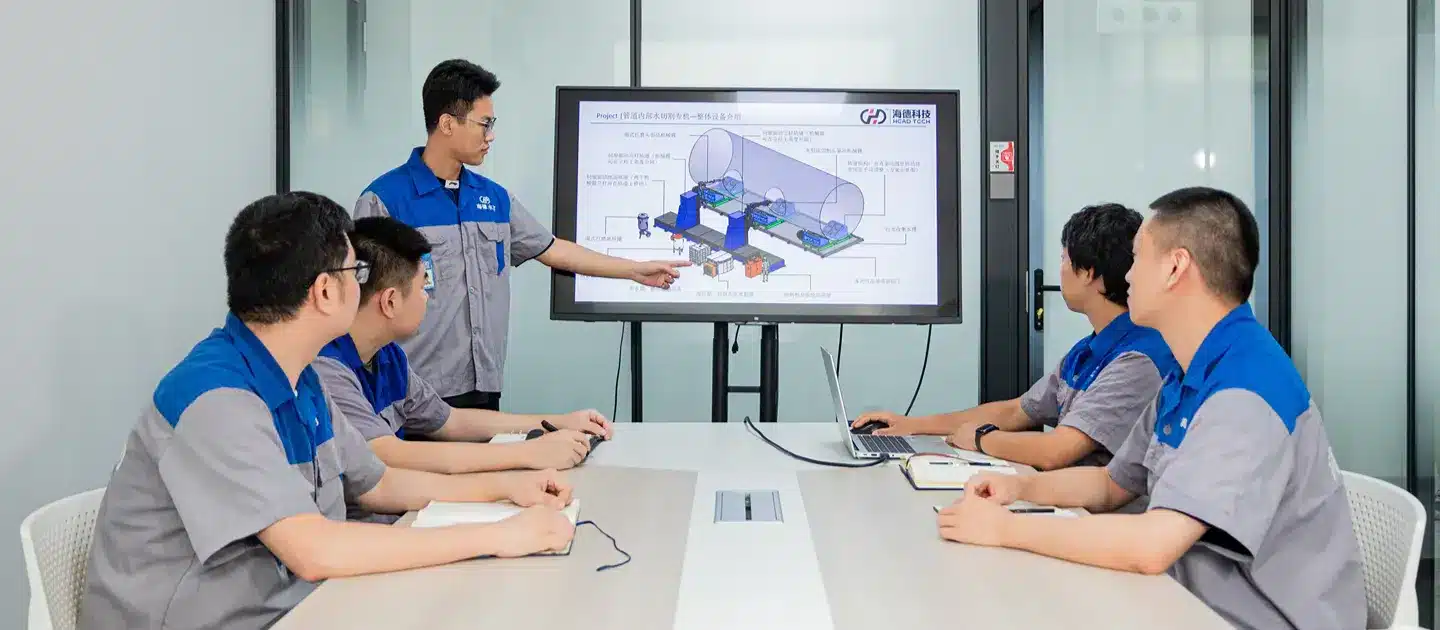 A group of R&D team members are gathered around a table for a meeting, discussing a technical diagram on a large screen.